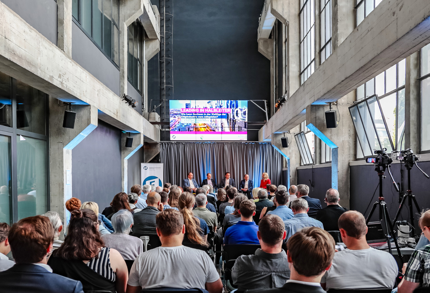 Foto: Publikum hört Podiumsdiskussion in hoher Industriehalle mit Leinwand