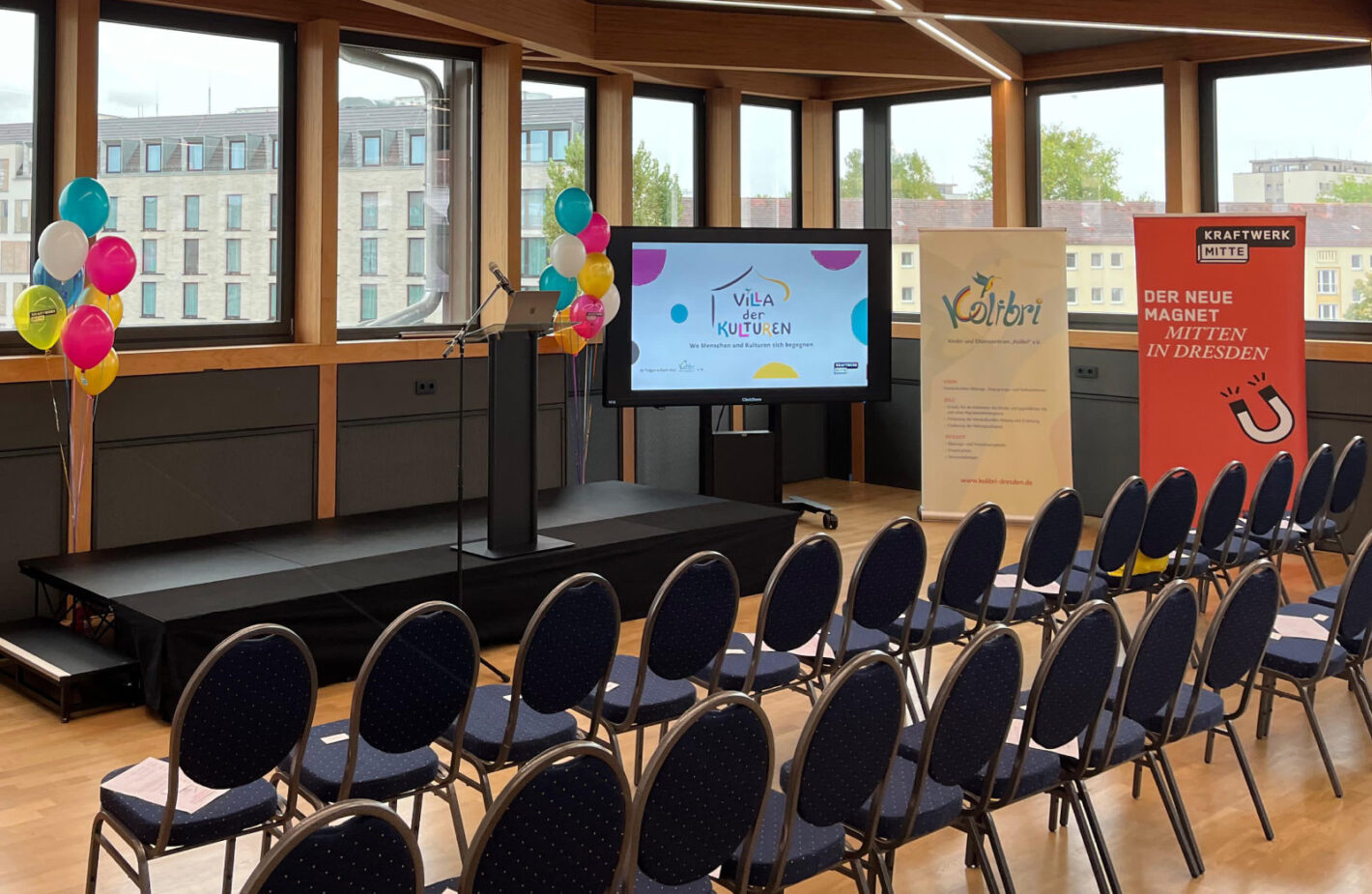 Foto: Veranstaltungsraum in der Villa mit Stühlen, Bühne, Mikrofon, Ballons und Monitor.