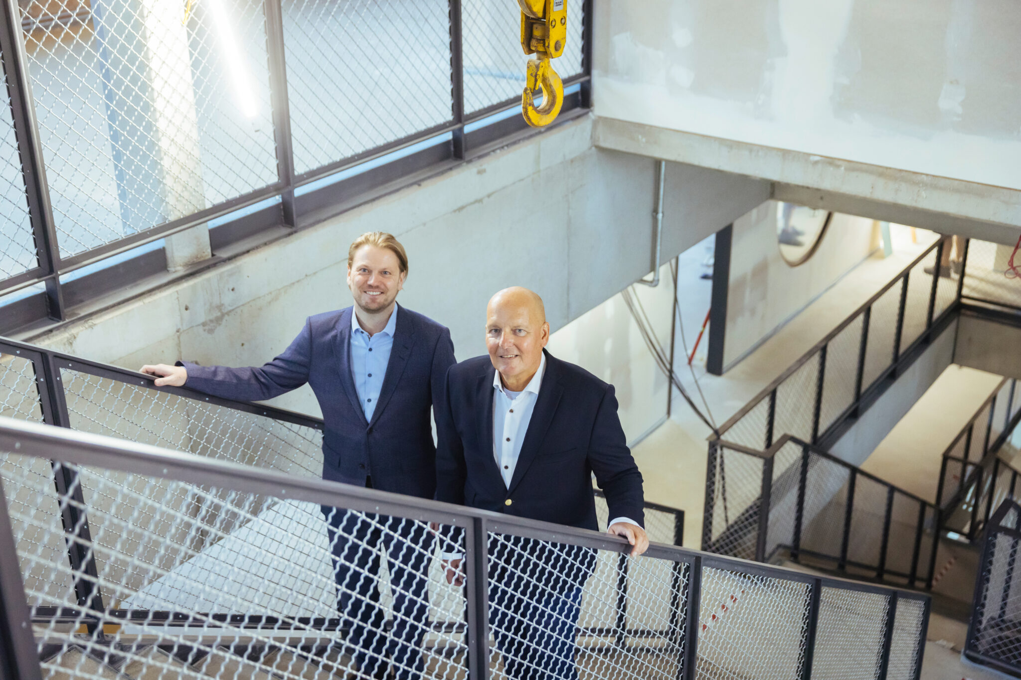 Foto: Zwei Männer im Anzug stehen lächelnd auf einer Treppe in einem modernen Rohbau.